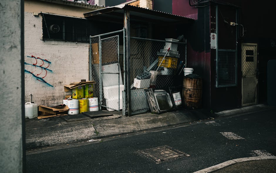 A small outdoor storage area located on the pavement in front of a building, enclosed by a metal chain-link fence with a corrugated metal roof. Inside the enclosure, various items are visible, includi